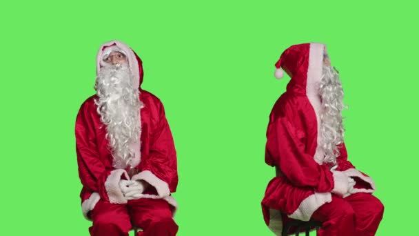 グリーンスクリーンの背景に座って椅子にサンタクロースの服を着た男 父のクリスマス実施 12月の季節の休日 伝統的なイベントを広告する若者 — ストック動画