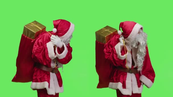 父のクリスマス実施形態は スタジオでプレゼントボックスとバッグを運び グリーンスクリーンに対する贈り物でサンタクロースのように振る舞います おもちゃの大きな象徴的な袋 休日のお祝いを持つ若い大人 — ストック動画