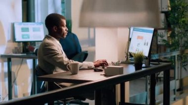 Afrika kökenli Amerikalı muhasebeci gece vardiyasında masa başında oturmuş, modern ofiste iş gelirleri istatistikleri üzerinde çalışırken, arka plandaki meslektaşı mola veriyor.