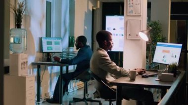 Ofiste iş arkadaşıyla gece vardiyasında çalışan mutlu Afro-Amerikan çalışanın portresi. Çalışma alanındaki titiz meslektaşlar, son teslim tarihinden önce takım projesi için bilgisayar görevleri yapıyorlar.