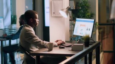 Afrika kökenli Amerikalı bilgisayar operatörünün portresi gece vardiyası sırasında masasında oturuyor, arka plandaki iş arkadaşı tuvaletteyken online finansal iş belgelerine bakıyor.