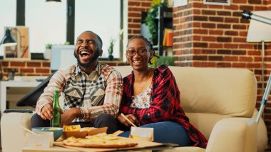 Smiling people laughing at comedy film on television, feeling happt spending time together. Young life partners enjoying movie on tv, eating chips and takeaway food. Tripod shot.