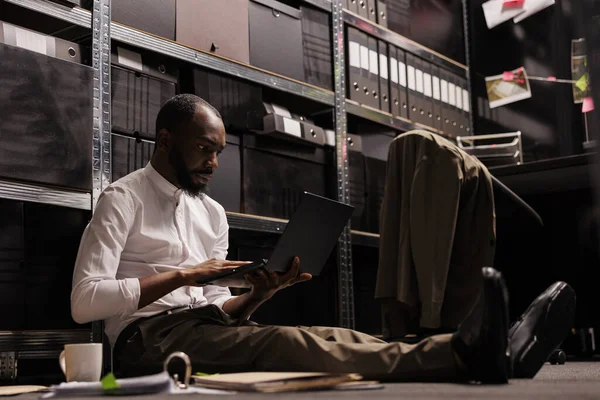 アフリカ系アメリカ人警官が床に座ってノートパソコンを使っている 犯罪事件ファイルを研究する犯罪学者 夜間にダークオフィスのポータブルコンピュータの情報をチェック — ストック写真