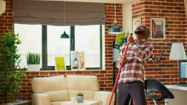 Cheerful man mopping apartment floors and listening to music, acting silly with dance moves at home. Young happy person doing spring cleaning with mop, having fun and smiling. Tripod shot.