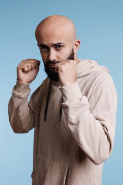 Arap adam boks pozisyonunda kameraya bakıyor, dövüşmeye hazır. Sıradan kapüşonlu giyen ciddi bir dövüşçü. Yumrukları sıkılmış ve öfkeli bir yüz ifadesi olan stüdyo resmiyle ayakta duruyor.