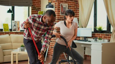 Diverse couple showing dance moves and having fun cleaning apartment, using mop to sweep dust and vacuum at home. Life partners dancing and singing, using washing solution, spring cleaning.
