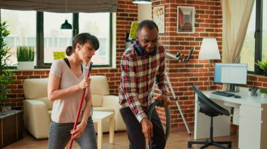 Diverse life partners dancing and sweeping dust off floors, using vacuum cleaner and washing solution. Cheerful couple laughing and enjoying spring cleaning in apartment, household chores.