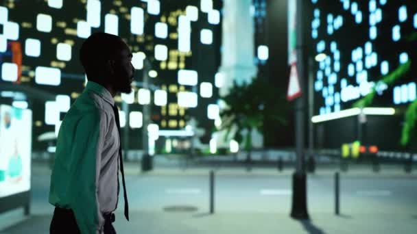 夜のアフリカ系アメリカ人は街灯の下を歩き 明るい歩道から近代的なオフィスビルを眺める 街中のナイトタイム遊歩道を楽しむ自信のある男 — ストック動画