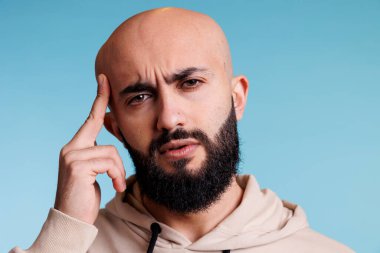 Arap adam migren krizi portresi çekerken tapınağı ovuyor. Genç, Arap, kel ve sakallı bir adam baş ağrısı çekerken yüzünü buruşturuyor ve kameraya bakıyor.