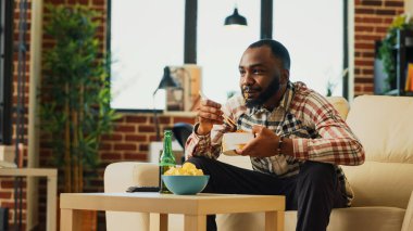 Happy guy eating noodles with chopsticks at home, enjoying asian food from delivery. Young adult relaxing with bottle of beer and takeaway meal, laughing at favorite tv show. Handheld shot.