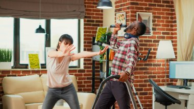 Interracial partners having fun cleaning room with vacuum cleaner and mop, dancing and listening to music. Modern cheerful people enjoying spring cleaning, vacuuming apartment floors.