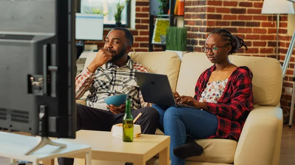 Young couple watching television together in living room, using laptop to browse internet and eating noodles with chopsticks. Life partners feeling relaxed at home with takeaway delivery.