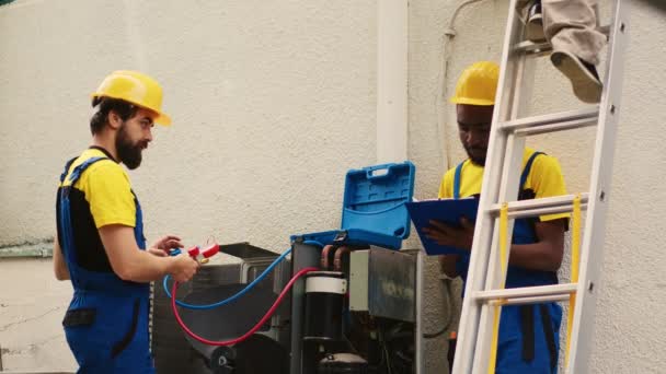 专家用压力指示器测量暖通空调系统中的氟利昂水平 而非洲裔美国同事在检查完屋顶冷凝器后从折叠梯子上下来 — 图库视频影像