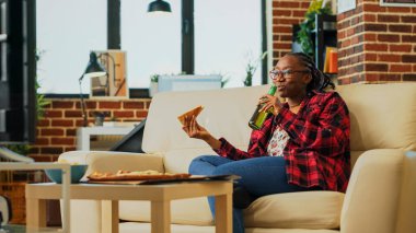 Female adult eating pizza slices from takeaway on sofa, enjoying film on television with alcohol bottles and fast food delivery meal. Cheerful girl having fun watching tv show. Handheld shot.