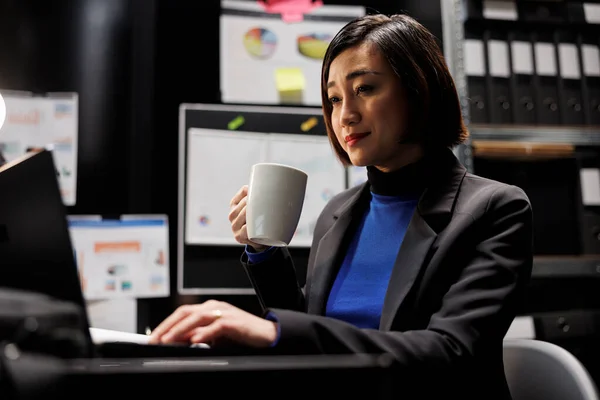 Cheerful bookkeeping businesswoman manager enjoying cup of coffee and imputing budget plan data on laptop in business office. Asian employee in file cabinet room with folders on bookshelves