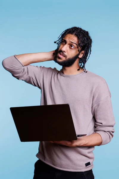 拿着笔记本电脑的阿拉伯程序员 对应用软件的开发没有把握 年轻人一边揉着头一边望着别处 一边在手提电脑上想着商业报告 — 图库照片