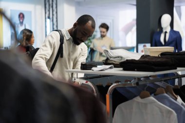 Afro-Amerikalı bir adam alışveriş merkezinin moda bölümünde alışveriş yaparken günlük kıyafetlerle rafları karıştırıyor. Butik mağazası müşterisi erkek giyim kuşamlarını incelerken moda kıyafetleri seçiyor.