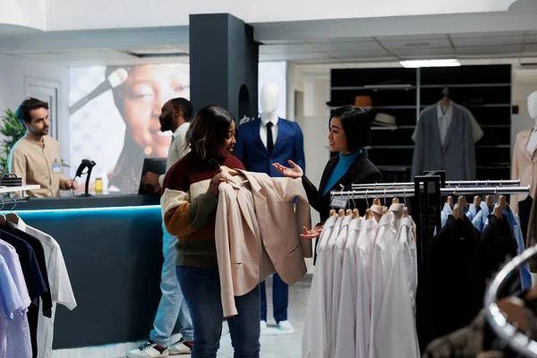 笑顔のアフリカ系アメリカ人女性がハンガーの上にジャケットを持ち ブティックでコンサルタントと話す 衣料品店で服を選択しながらファッションのトレンドを議論する顧客とアシスタント — ストック写真