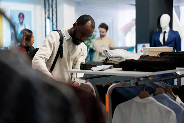 アフリカ系アメリカ人男性は ショッピングモールのファッション部門でカジュアルな服でラックをブラウズします トレンディな服を選択しながらメンズウェアのアパレルを検討するブティックストアの顧客 — ストック写真