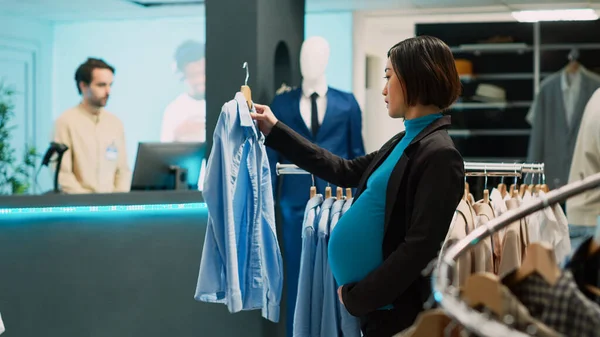 妊娠中の顧客はハンガーの流行の服を分析し 将来のお母さんは割引で購入したい 若いです女性で赤ちゃんバンプチェックファッショナブルな商品で衣料品店 中小企業 — ストック写真
