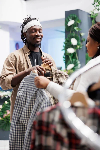 Two friends browsing clothes on hangers and choosing plaid shirt in shopping mall. African american girlfriend helping boyfriend choosing trendy casual outfit in clothing store