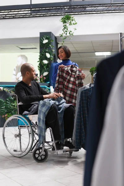 衣料品店 アジアの女性の顧客は 物理的な障害を持つアラブ人男性にハンガーに配置されたシャツを示しています 車椅子のモールブティッククライアントは アパレルショッピング中に売り手から支援を受ける — ストック写真