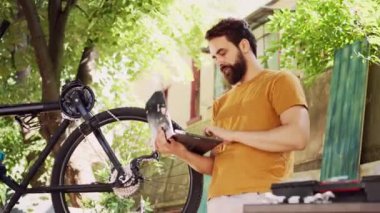 İnternet ve teknoloji kullanarak bisiklet ekipmanlarını koruyan ve koruyan genç sporcu. Ev bahçesinde bisiklet tamir eden laptoplu, spor düşkünü, beyaz bir erkek..