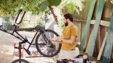 Erkek bisikletçi kişisel bilgisayar tutuyor ve bisiklet pedalını ve kolundaki hasarı kontrol ediyor. Aktif Kafkasyalı adam internette gezinmeyi, modern bisikleti tamir etmeyi ve korumayı seviyor.