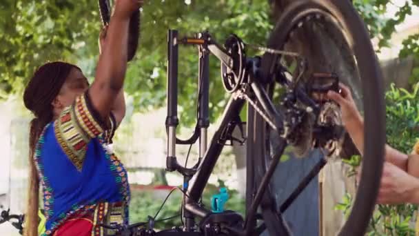 専門装置を使用して維持のための損傷した自転車を点検する適合および専用の多人種間ペア 自転車のペダルを修理し 壊れた車輪を取り除いているガールフレンドと話すボーイフレンド — ストック動画