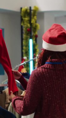 Dikey video Afrikalı Amerikalı perakende asistanı Noel Baba şapkası takıyor Asyalı müşterinin kış tatilinde Noel alışverişi mağazasında şenlikli kıyafetleri karıştırmasına yardım ediyor. Çalışan asistanı.
