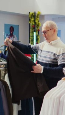 Dikey video yaşlı müşteri elbise raflarını karıştırıyor, Noel tatilinde aile üyelerine hediye olarak şık bir blazer ceket almak istiyor. İdeal bir hediye arayan yaşlı bir müşteri.