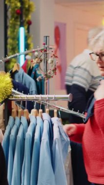 Dikey video sunucusu kadın Noel süslü giyim mağazasında spor ceket raflarını karıştırırken kocası arka plandaki tezgahta para ödüyor. Müşteriler kış tatilinde Xmas alışverişi yapıyor.