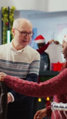 Dikey video yaşlı müşterisine, şık bir spor ceketin uygun olup olmadığına karar vermek için festival dekoratif giyim mağazasında perakende asistanı yardım ediyor. Çalışan Noel süslemesinde yaşlı adama yardım ediyor