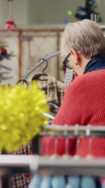Dikey video kasetteki yaşlı kadın giyim mağazası erkek bölümünde spor ceket raflarını karıştırıyor ve kış tatili sezonunda koca için mükemmel bir Noel hediyesi arıyor. Müşteri verecek kıyafet arıyor.