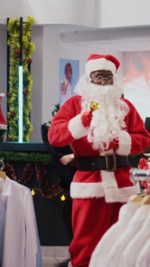 Noel Baba kılığındaki Noel Baba 'nın alışveriş merkezindeki giyim mağazasında çekilen dikey video Dolly. Denetmen Noel çanları çalıyor ve tatil temalı müşterileri selamlıyor.