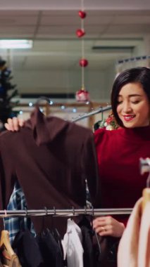 Dikey Videocu Kadın Noel giysi mağazasında kıyafetlerini karıştırıyor. Yeni yıl partisi için mükemmel kıyafetler arıyor. Moda mağazasının müşterisi ideal kıyafet arıyor.