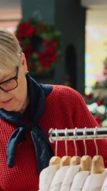 Dikey videolu yaşlı kadın kış tatili boyunca kostüm mağazasında kıyafetleri karıştırıyor. Alışveriş merkezindeki Noel dekore edilmiş moda butiklerinde alışveriş çılgınlığında kıdemli müşteri.