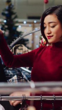 Dikey videolu Asyalı müşteri Noel temalı mağazada kıyafet raflarını karıştırıyor, bluz ölçülerine uyuyor mu diye kontrol ediyor. Bayram sezonunda Xmas 'lı bir kadın moda butiği süslüyor.