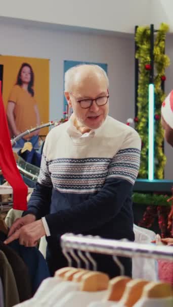 クリスマスの垂直ビデオ熟練した従業員は 高級ブレーザーを金持ちのシニアクライアントに混乱させ 質の高い素材で巧みに作られた衣服を彼に納得させます — ストック動画