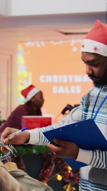 圣诞主题服装店的垂直视频管理器 戴着圣诞帽 手持剪贴板浏览衣架 在节假日促销促销活动中整理价格标签 — 图库视频影像