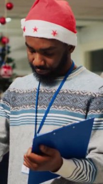 Dikey Afrikalı Amerikalı çalışan Noel süslemeli moda mağazasında elbise raflarını karıştırıyor, ayarlanmış fiyatları panoya yazıyor. İşçi ceketleri inceliyor, hasar arıyor.