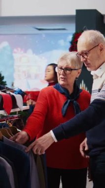 Dikey video kasedi yaşlı karı koca Noel alışverişi sırasında kostüm mağazasında elbise raflarına göz atıyorlar. Resmi kıyafetler almak isteyen yaşlı müşteriler, tatillerin tadını çıkarıyorlar.