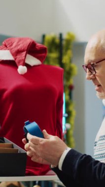Dikey video Yaşlı Adam kış tatili sezonunda Noel süslemeli mağazada zarif moda kemer koleksiyonuna göz atıyor. Üst düzey müşteri, yeğenlerine hediye almak istiyor.