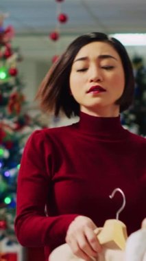 Noel süsleri mağazasında kış tatili boyunca zarif ceketlerin arasında gezinen mutlu bir kadının dikey video portresi. Gülümseyen müşteri moda ürünlerini kontrol ediyor