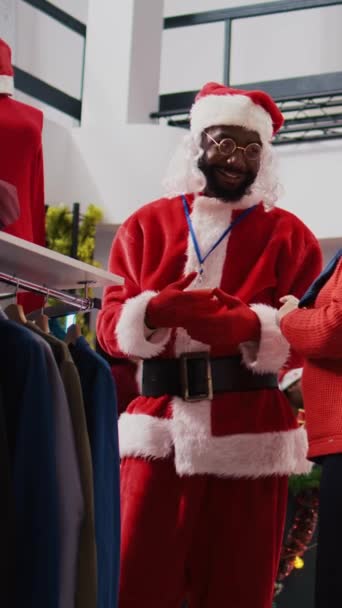 サンタクロースとして服を着た垂直ビデオ従業員は 装飾されたクリスマスの家族のためのギフトを購入しようとしている高齢者クライアントに赤い衣料品に関する資料と価格情報を提示します — ストック動画