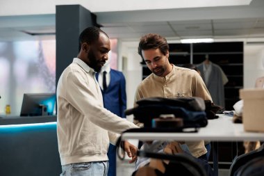 Giyim mağazası müşterisi ve asistanı giyim mağazalarını keşfederken moda trendleri hakkında konuşuyorlar. Afrika kökenli Amerikalı bir adam butik işçisinden kıyafet seçiminde yardım istiyor.