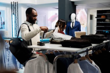 Müşteri, kıyafet mağazasından alışveriş yaparken kravat ve gömlek eşleşmesi yapıyor. Afro-Amerikalı adam resmi kıyafet ve aksesuar seçiyor, galeride giysi ölçülerini ve tarzlarını kontrol ediyor.