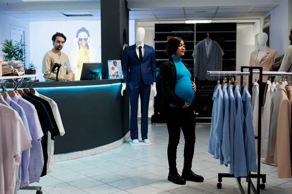 ショッピングモールのファッションブティックで母性服を探している若い妊娠中のアジアの女性 ショッピングセンターの掛かるアパレルでラックの近くに立っている期待のお母さん 衣服を選ぶ — ストック写真