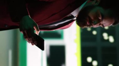 Dikey video klas iş adamı gece yürüyüşünde mesajları kontrol ediyor, şehir merkezinde geziniyor ve kaldırımda yol tarifi arıyor. Kendine güvenen bir adam şehirde yürüyor, akşam gezintisi yapıyor.