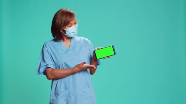 Hemşire krom anahtar yeşil ekranda açıklayıcı bir sağlık bandı sunuyor. Hastane çalışanı telefonu peyzaj modunda tutuyor, video gösteriyor, mavi stüdyo arka planında izole edilmiş.